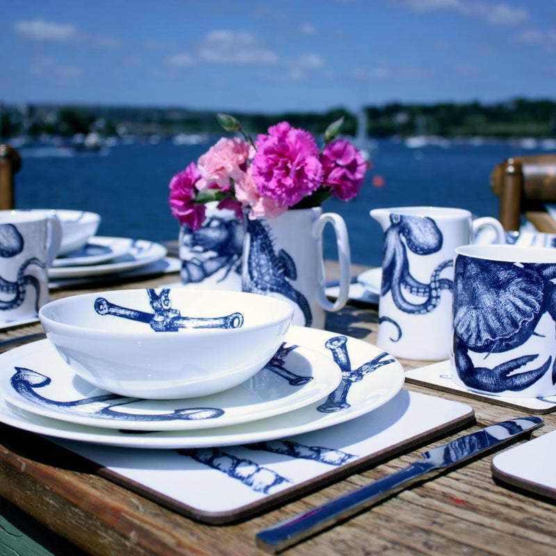 Bone china white mug featuring hand drawn Crab design in classic Navy at a table setting.Pink carnations in a Seahorse Jug surrounded by our other classic designs with harbour view in the background
