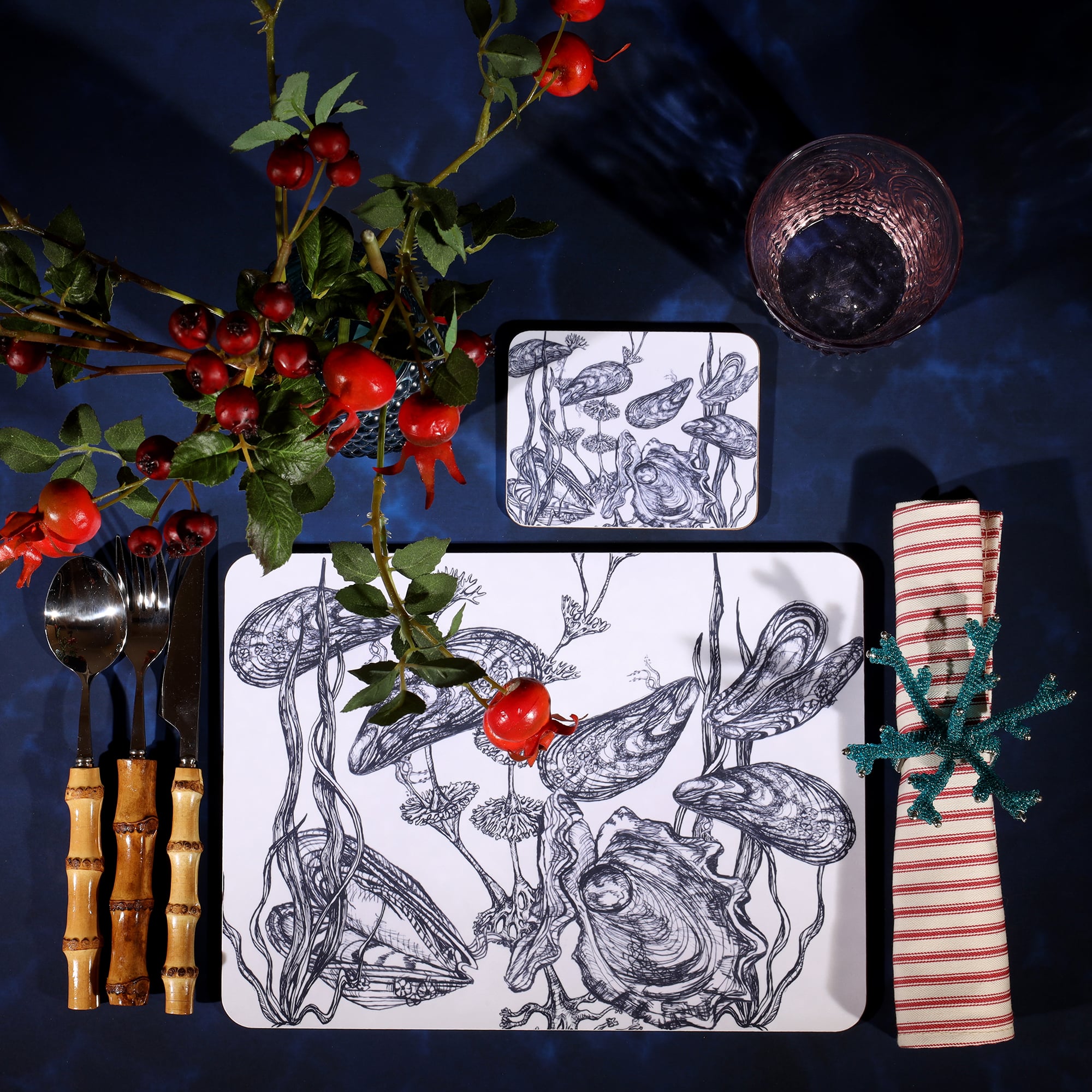 Mussel and Oyster  Design in Navy on a white Coaster with a matching Placemat in a table setting on a dark table cloth.On the table is bamboo cutlery, stripe napkin and a glass with rosehips.