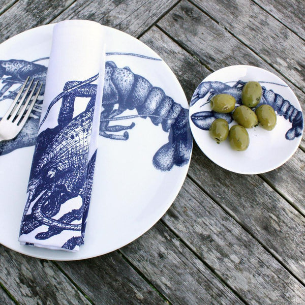 Nibbles bowl in Bone China in our Classic range in Navy and white in the Lobster design next to a matching plate on a wooden table.On the plate is a folded napkin