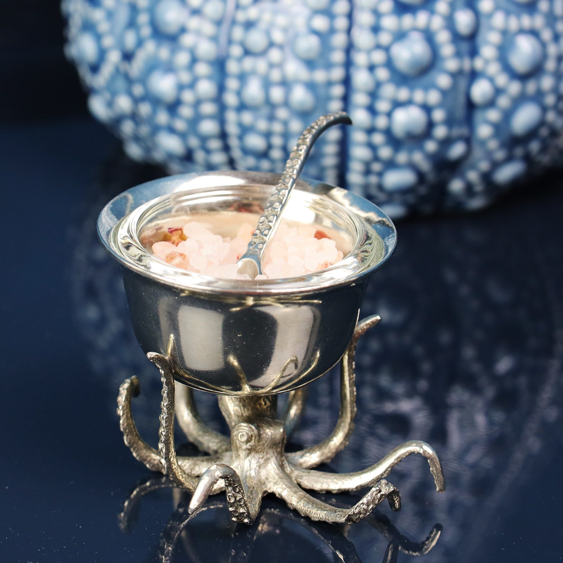 Pewter Octopus Condiment Bowl with the spoon containing salt,sits on a table with other pewter items and a jug in the background