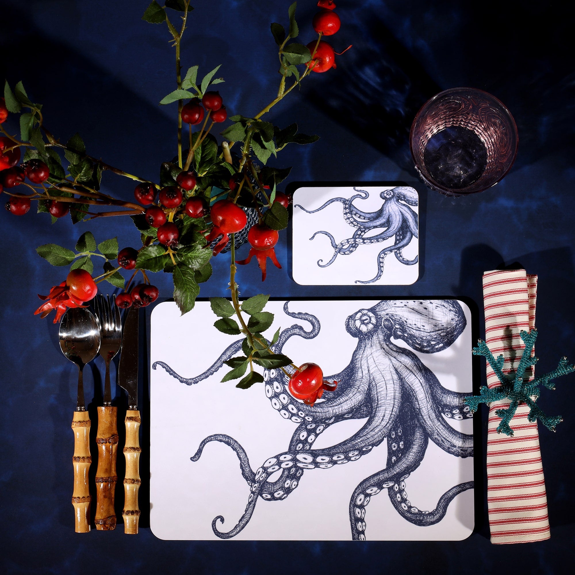 Octopus Design in Navy on a white Coaster with a matching Placemat in a table setting on a dark table cloth.On the table is bamboo cutlery, stripe napkin and a glass with rosehips.