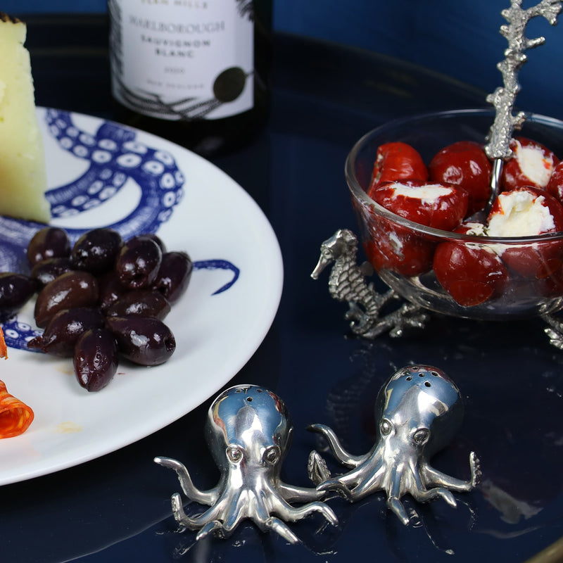 Pewter Octopus Salt and Pepper Shakers placed on a table with a plate with Olives on,next to that is a glass bowl and a bottle of wine in the background