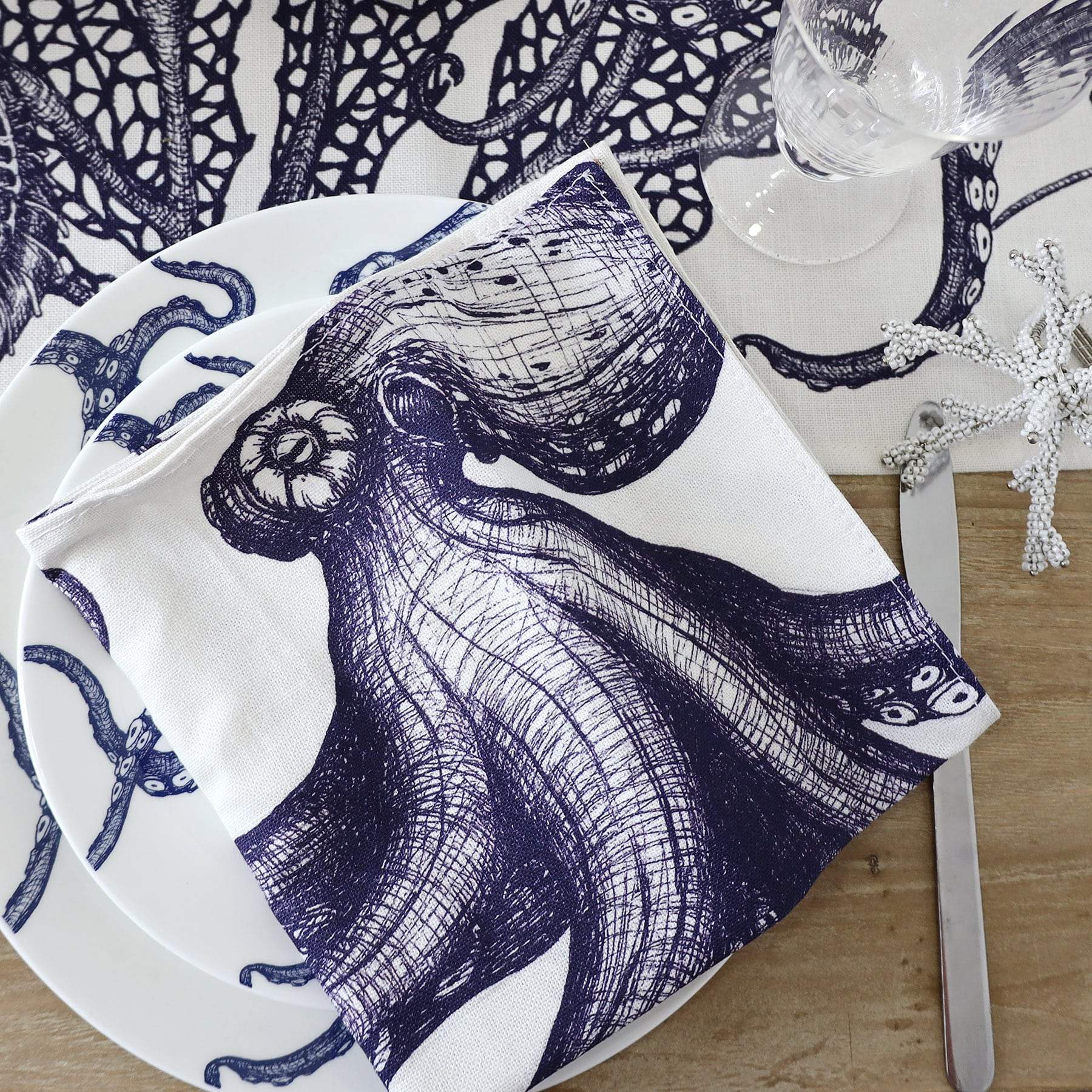 Blue & white linen napkins with seahorse, lobster, octopus and jellyfish hand drawn illustrations,set of four,close up of the Octopus one placed on an octopus dinner plate.All placed on a sea creature tablecloth