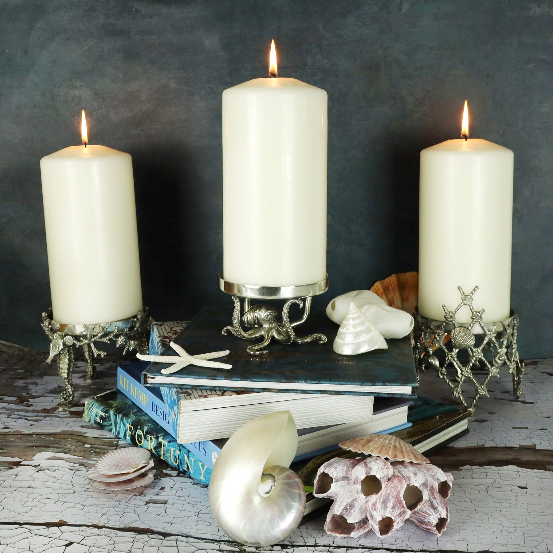 Close up shot of three large Pewter Candle Holder featuring intricate seahorses as the base placed on a table with books and shells on the table