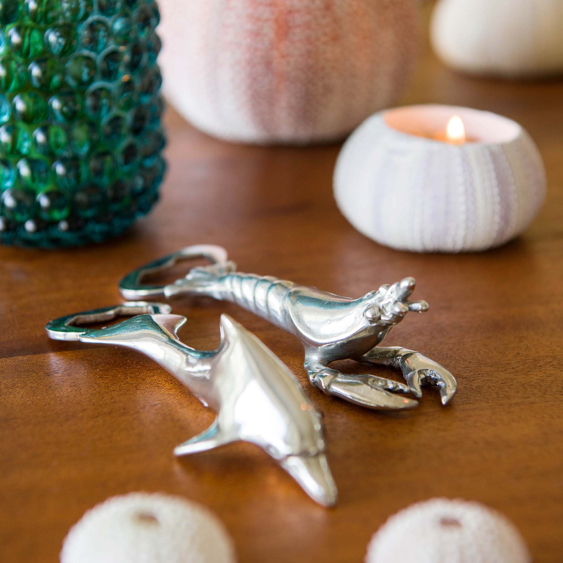 Pewter Dolphin and a Lobster shaped Bottle Opener placed on a table in front of a shell candle and candle holders in the background