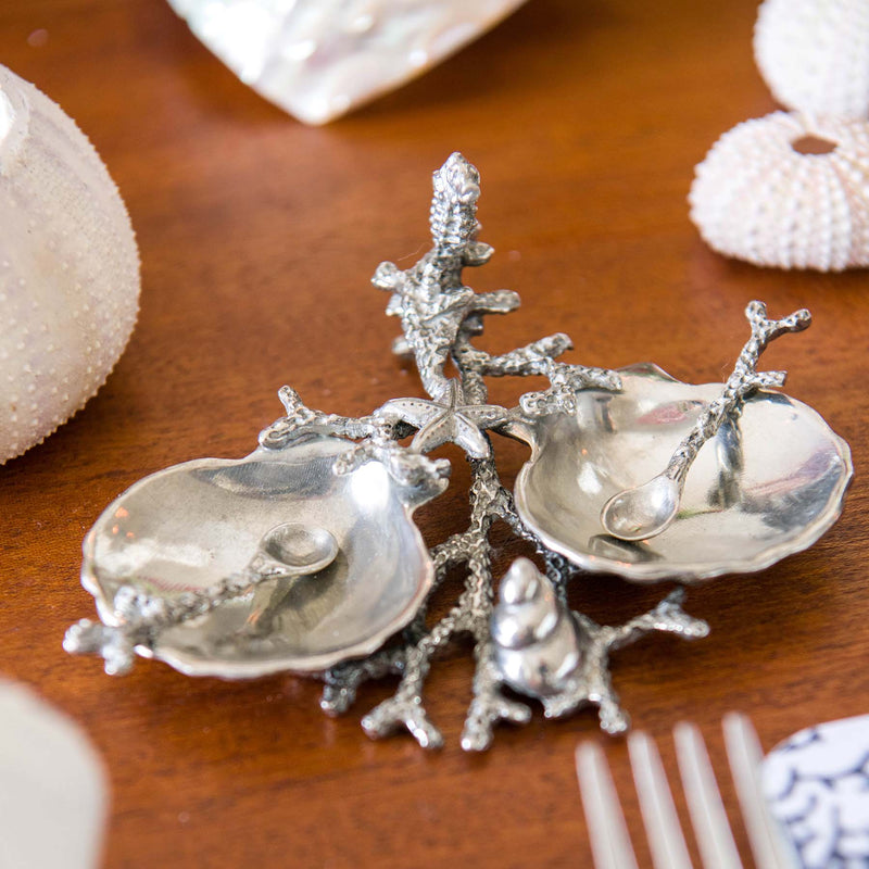 Pewter Shell & Coral Condiment Set consisting of two pewter scallop bowls held together by a sea coral design pewter structure.It has a starfish, tiny seahorse  and a shell on the branches,placed on a table with other shell items