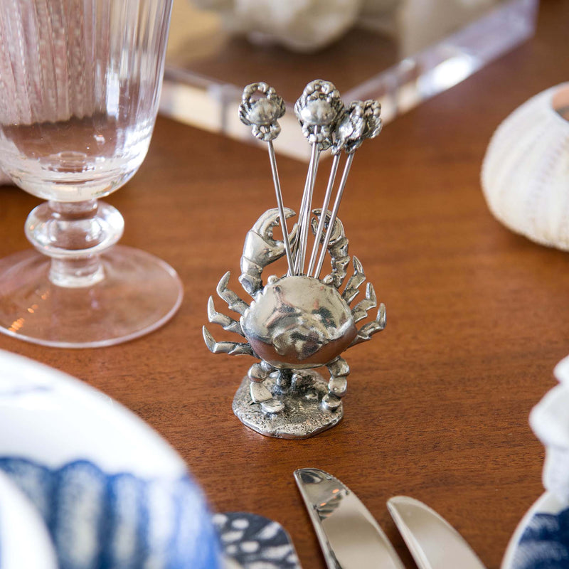 Pewter Crab shaped Pick Set with crabs at one end of each of the picks placed on a table ,a glass and tableware can be seen surrounding it