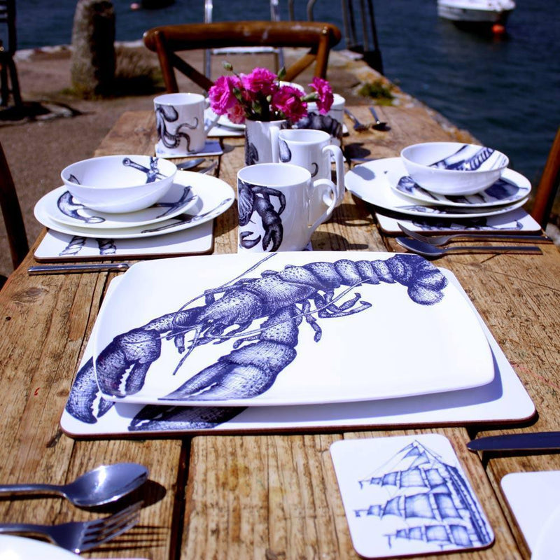 Platter in Bone China in our Classic range in Navy and white in the Lobster design on a outdoor wooden table placed on a tablemat.In the background are other place settings in our classic designs with flowers on the table and you can see the sea in the background