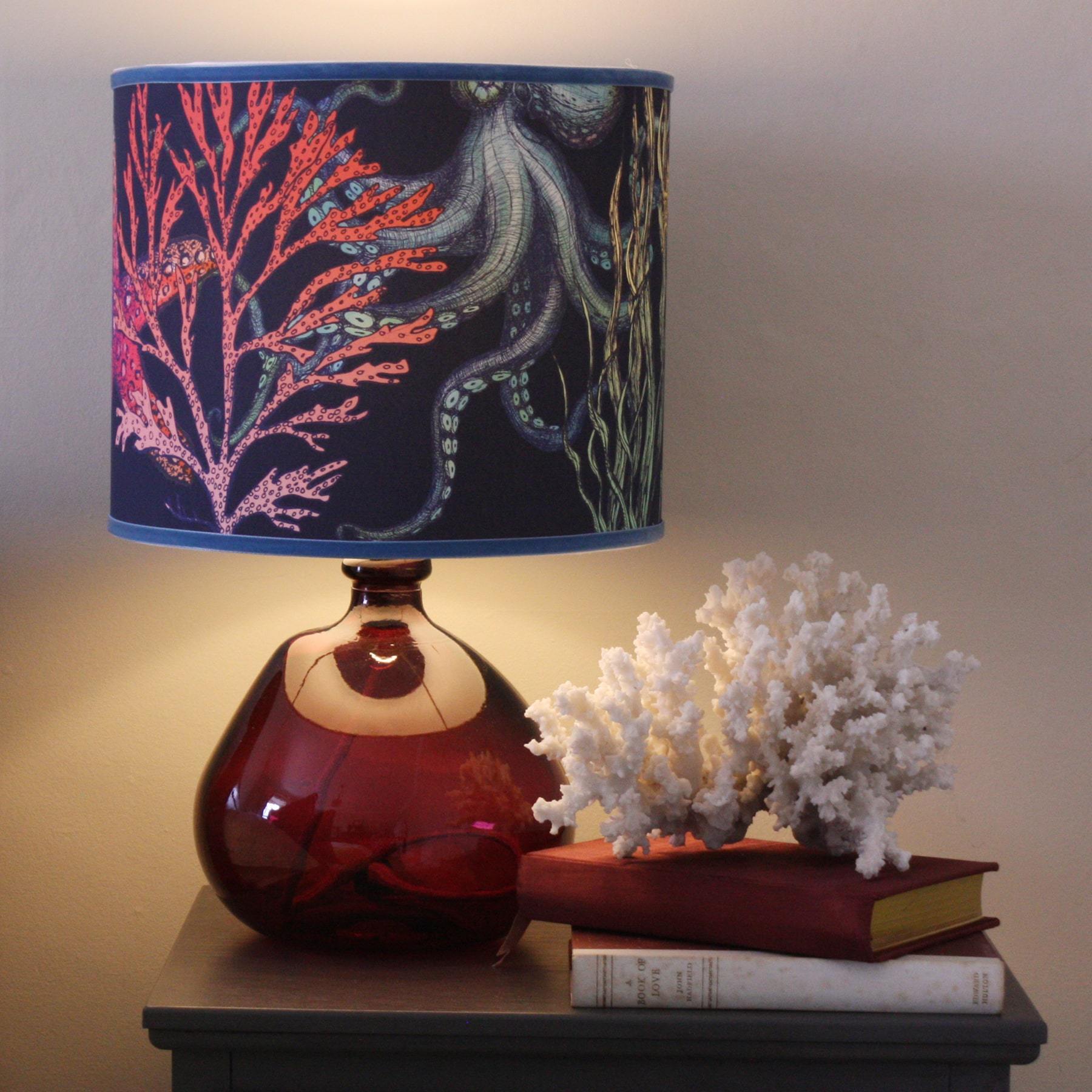 Rainbow Reef Navy Shade With Octopus,Seahorse,Starfish and Seaweed Design in bright colours on a pink lampbase on a sideboard with a couple of books and a coral sculpture.
