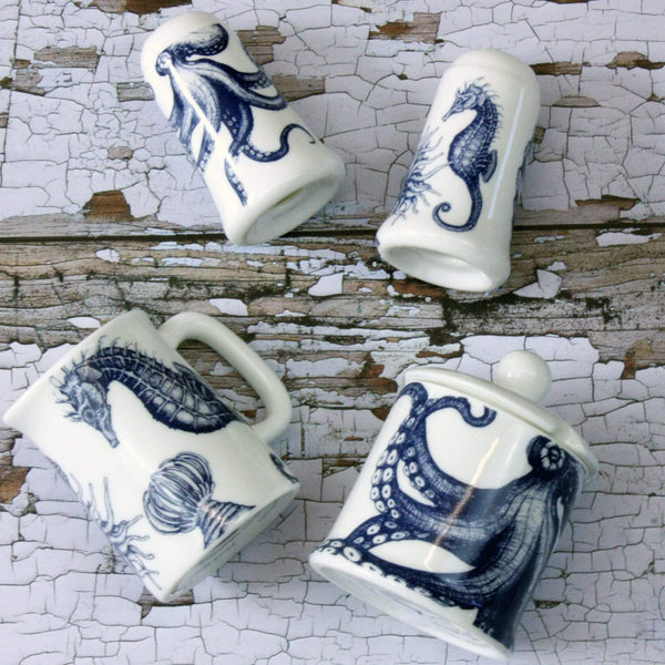 Bone china salt an pepper pots in our classic navy and white.Salt has seahorse, shell and an anenome on and the pepper has an octopus.They are both lying on a distressed wooden table also lying on the table is a seahorse jug and an octopus jam pot, all seen from an aerial view