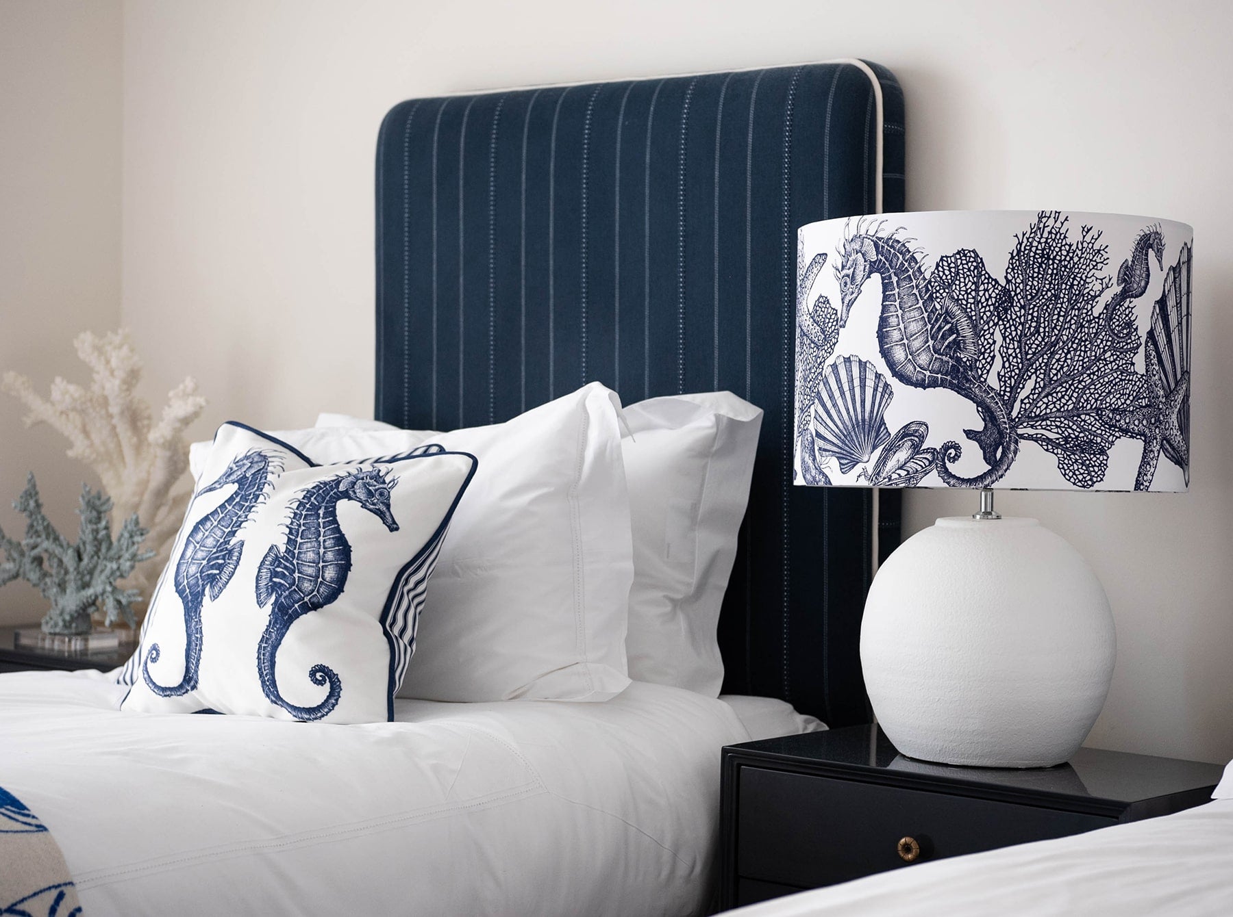 Our Classic Navy Seahorse design on a white background on a white lampbase on a side table between two single beds.On one of the beds you can see our classic cushions with a seahorse design.