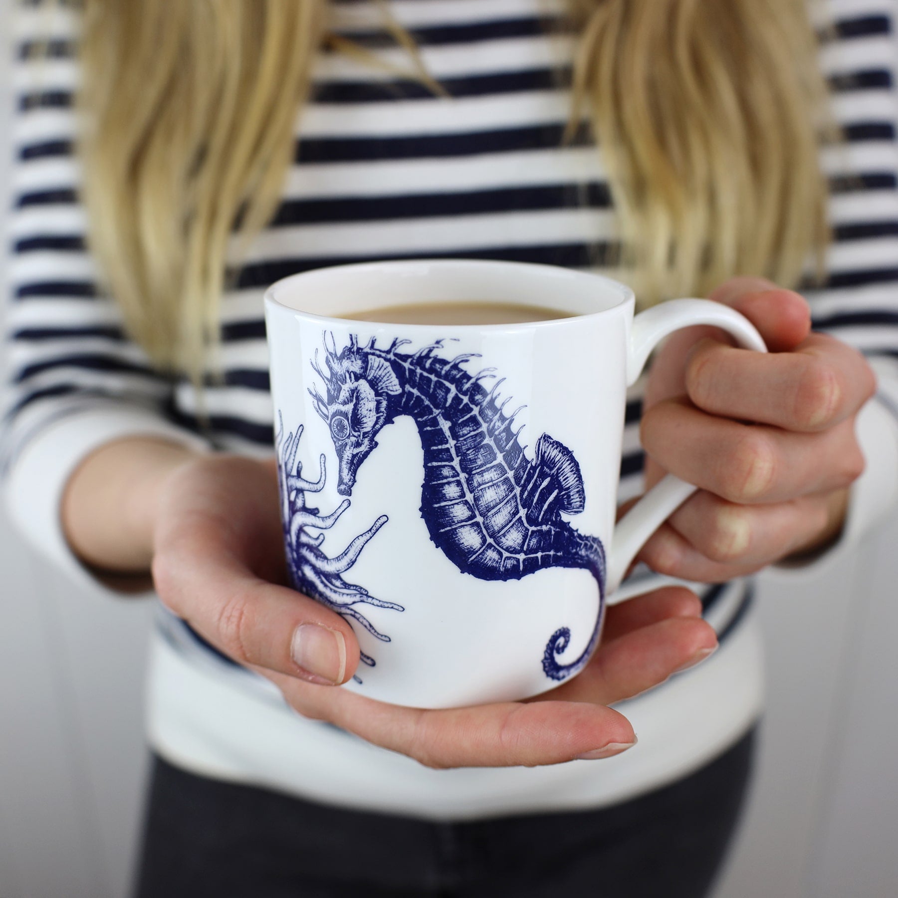 The photo shows Bone china white mug featuring hand drawn seahorse,scallop shell and anemone design in classic Navy held by a woman