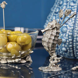 Pewter Seahorse shaped Pick Set with seahorses at one end of each of the picks placed on a table in front of an urchin Jug and a condiment glass bowl containing olives