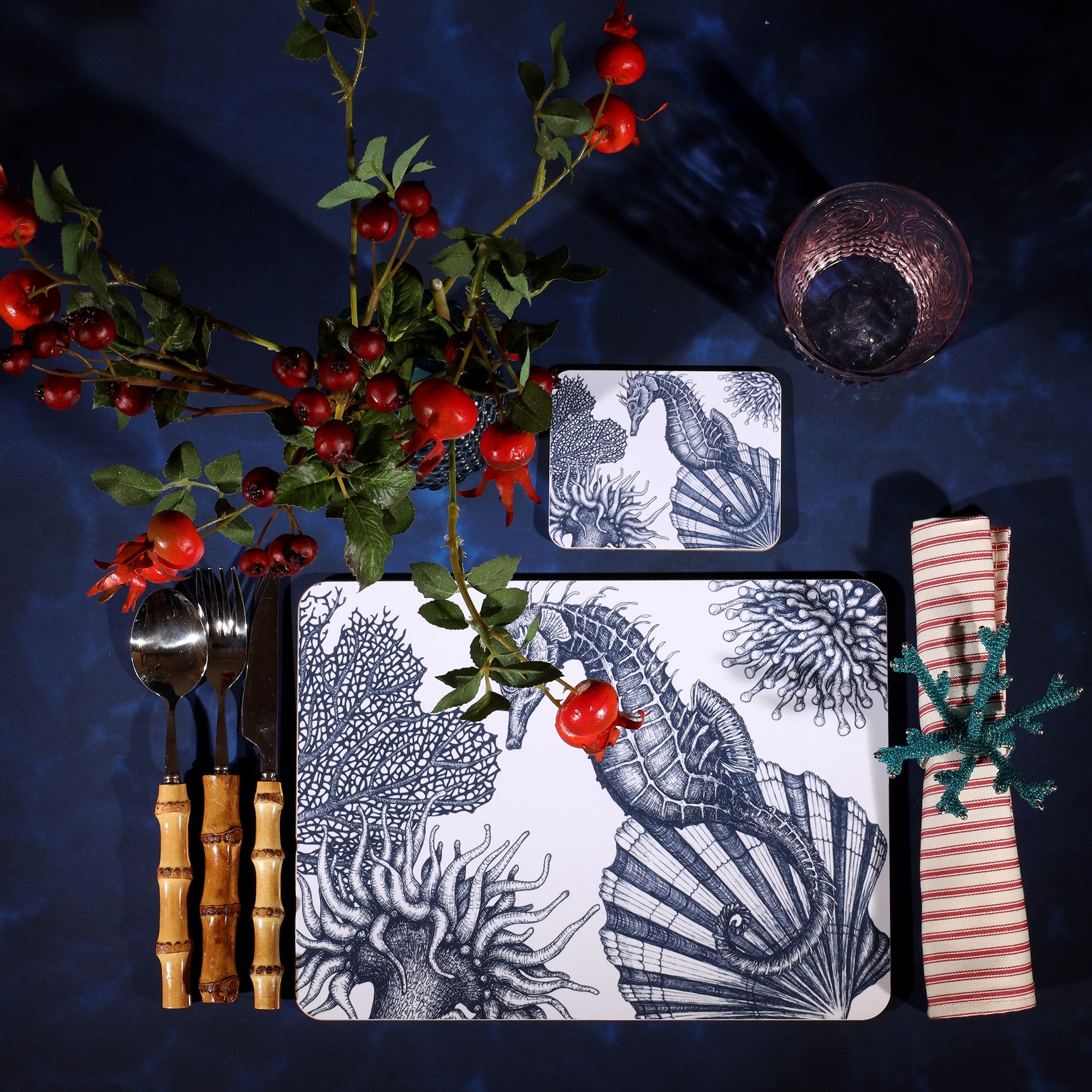 Seahorse Design in Navy on a white Coaster with a matching Placemat in a table setting on a dark table cloth.On the table is bamboo cutlery, stripe napkin and a glass with rosehips.