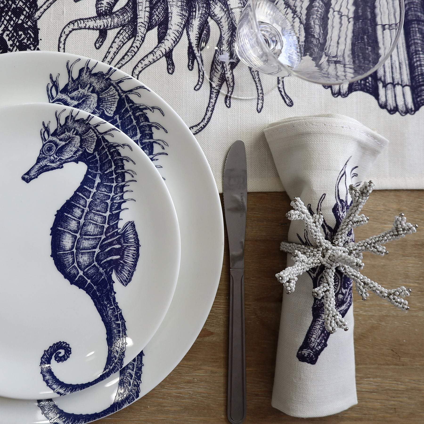 Blue & white linen napkins with seahorse, lobster, octopus and jellyfish.Close up of the Seahorse displayed next to a seahorse dinner and side plate.Also on the table is one of our table runners 
