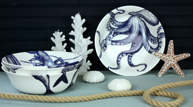 Serving bowl in Bone China in our Classic range in Navy and white in the Octopus design placed on its side on a table,next to a pile of various serving bowls.On the table are candles shaped as Coral and a starfish and some shells,a rope winds through the items