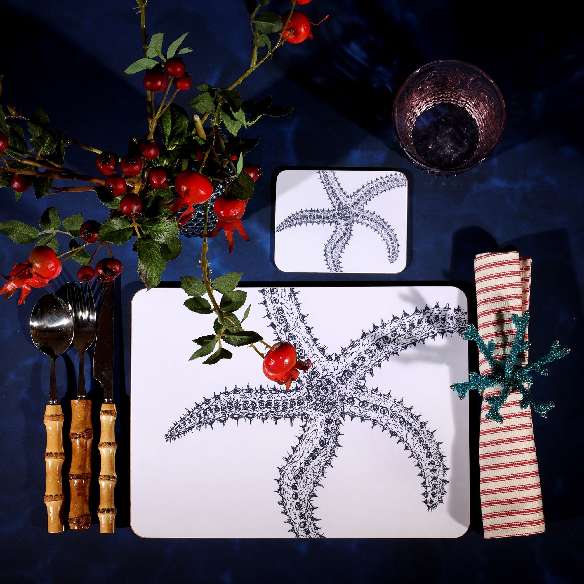 Starfish Design in Navy on a white Coaster with a matching Placemat in a table setting on a dark table cloth.On the table is bamboo cutlery, stripe napkin and a glass with rosehips.