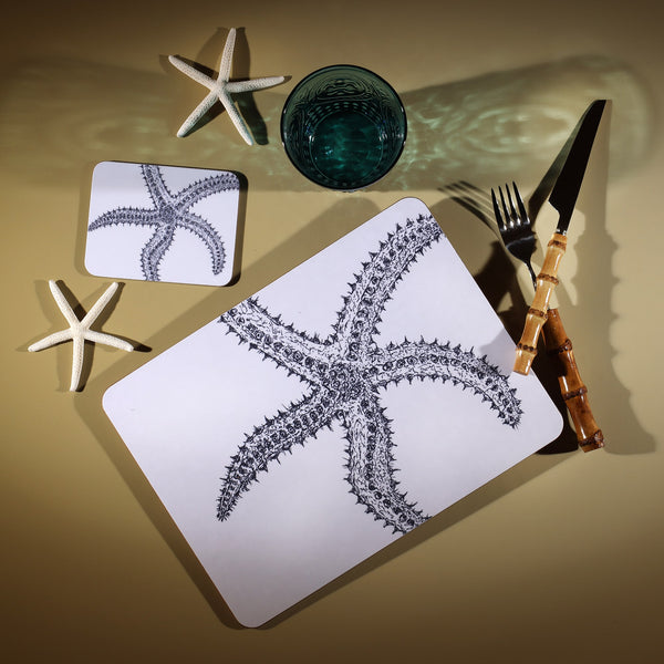 Starfish Design in Navy on a white Coaster with a matching Placemat.On the table is a bamboo cutlery set and a blue coloured glass