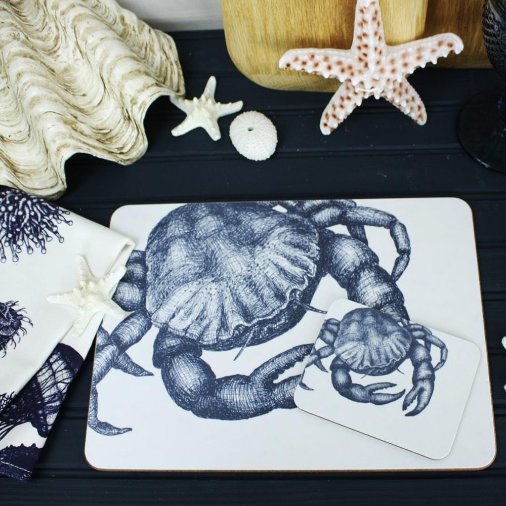 Blue and White Placemat with our Crab design with matching Coaster placed on a navy table.The table is also decorated with shells and table linens