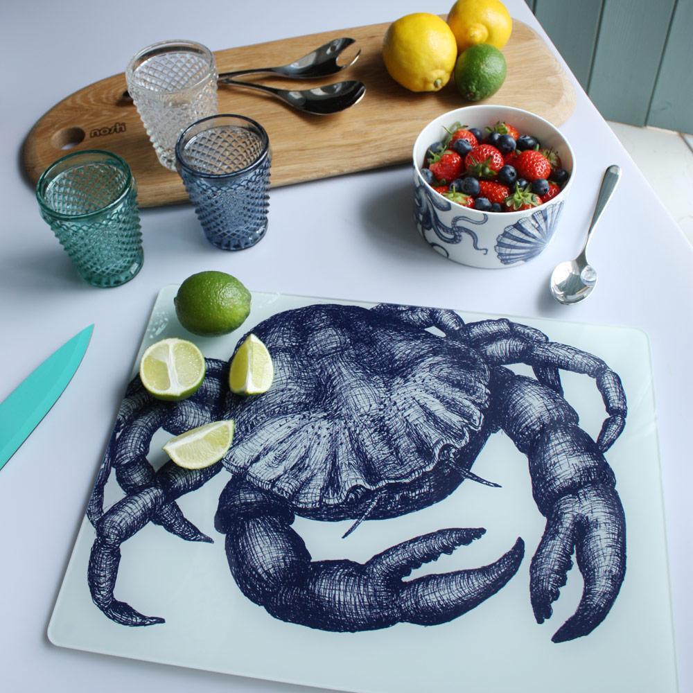 Worktop saver on toughened recycled glass with our Crab design placed on a table with coloured glasses,a wooden board,fruits and a bowl on the table