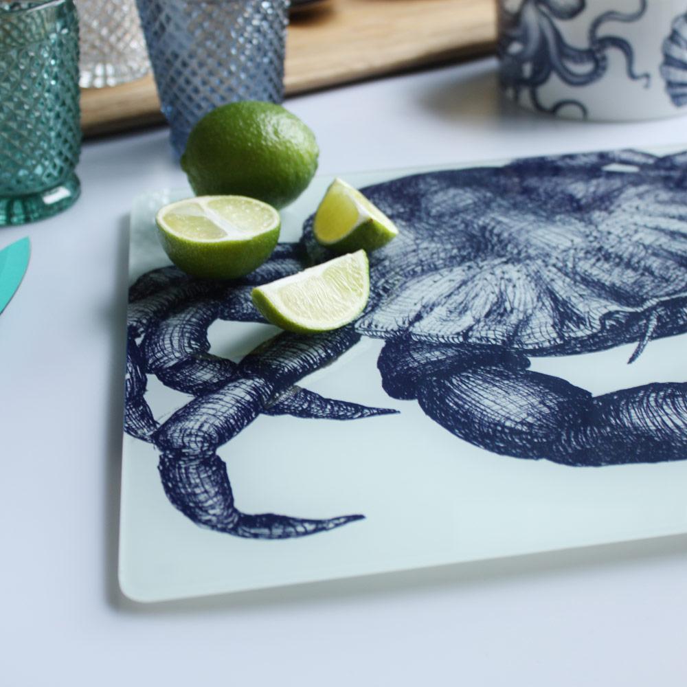 Close up of Worktop saver on toughened recycled glass with our Crab design placed on a table with coloured glasses,a wooden board,fruits and a bowl on the table