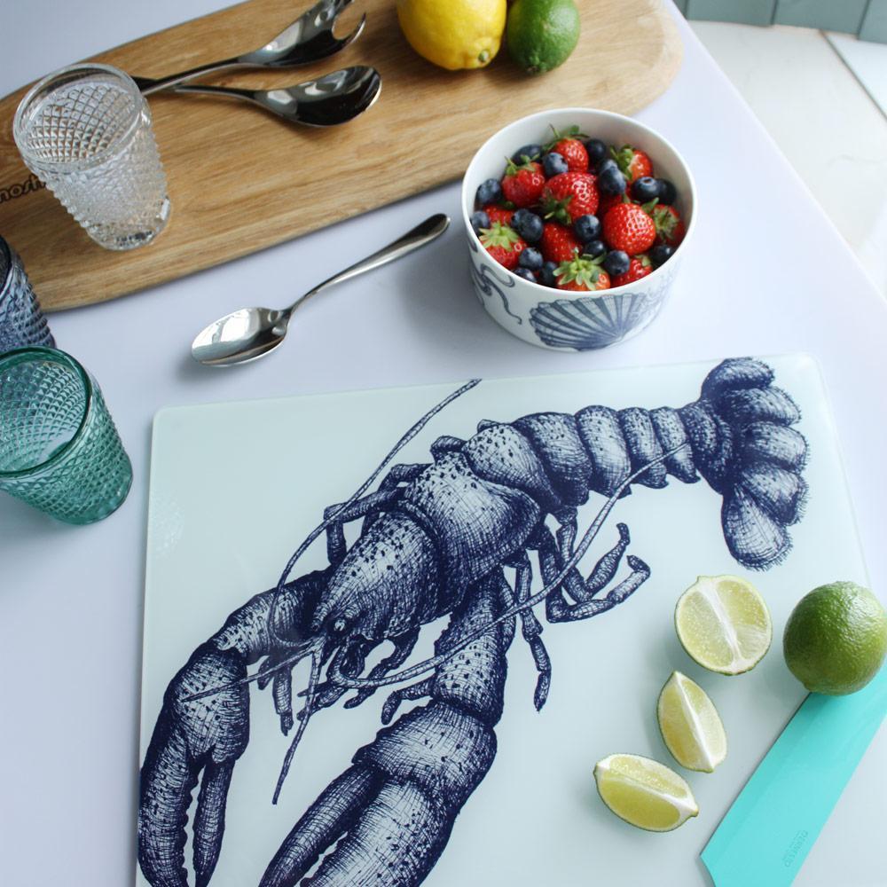 Worktop saver on toughened recycled glass with our Lobster design placed on a table with coloured glasses,a wooden board,fruits and a bowl on the table