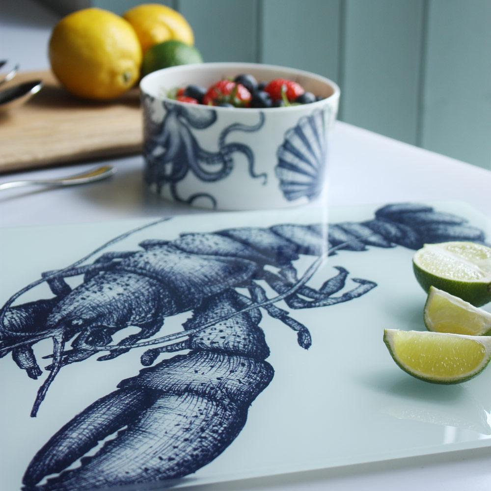 Close up of Worktop saver on toughened recycled glass with our Lobster design placed on a table with coloured glasses,a wooden board,fruits and a bowl on the table