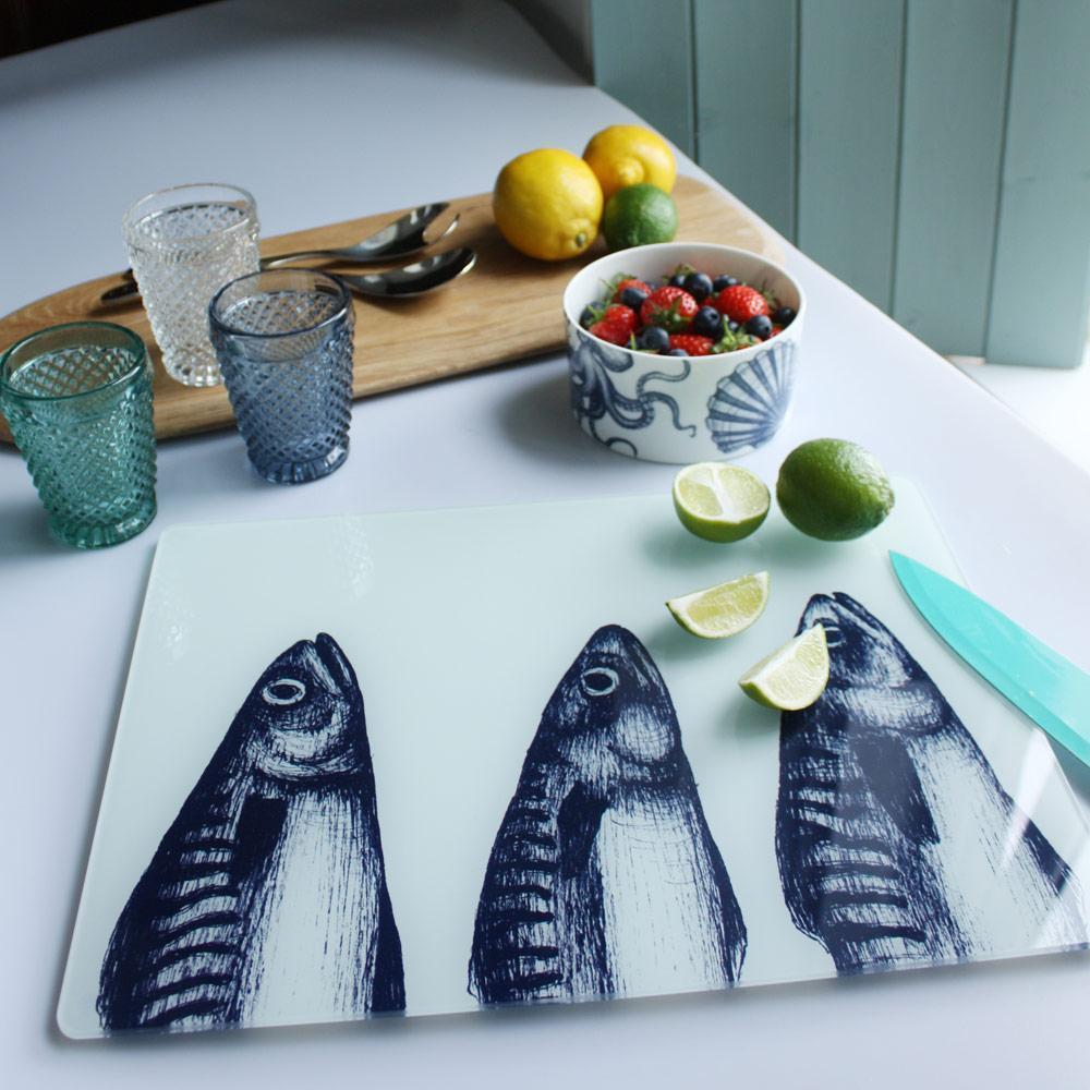 Worktop saver on toughened recycled glass with our Mackerel Heads design placed on a table with coloured glasses,a wooden board,fruits and a bowl on the table